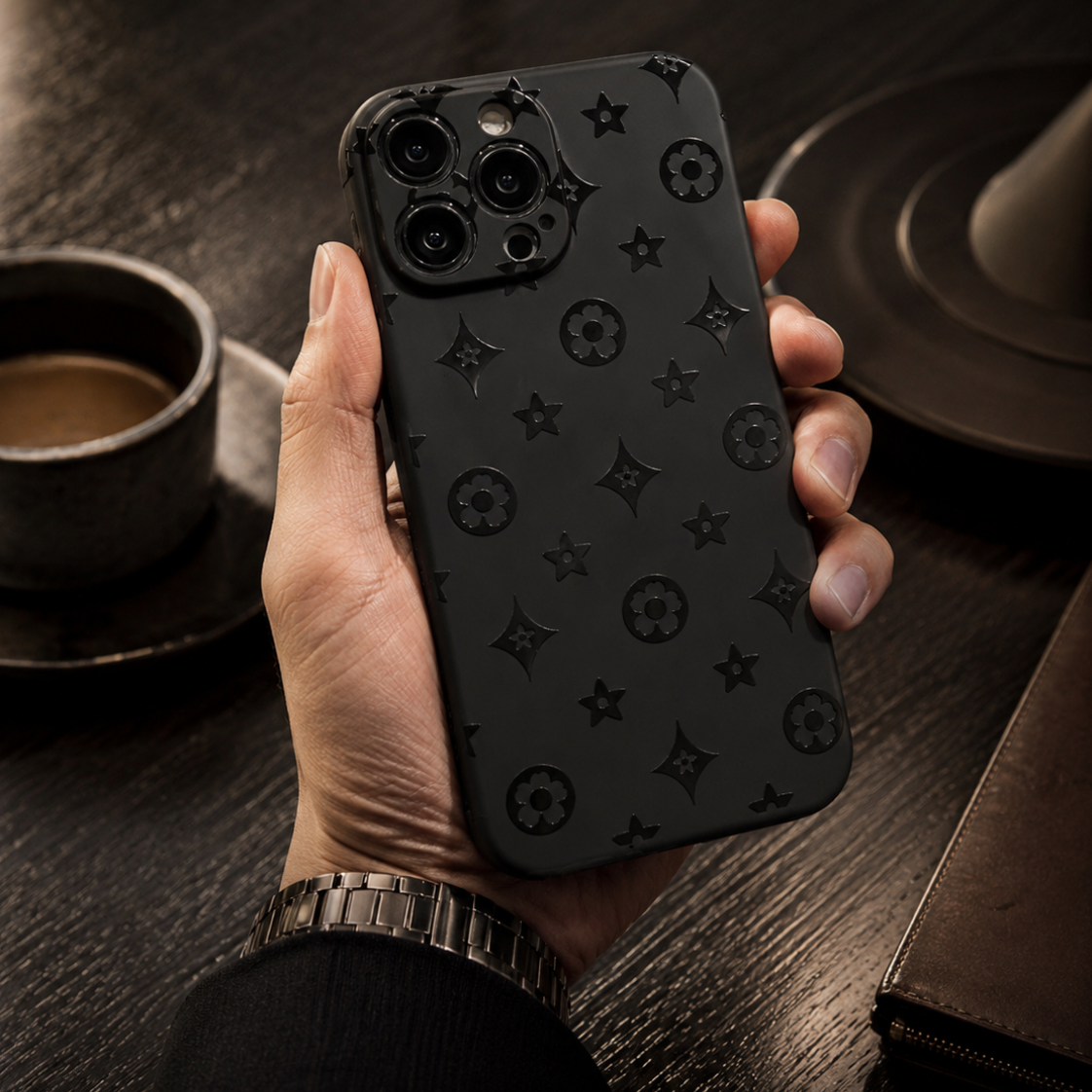 Hand holding a black phone case with star pattern on a wooden table with coffee cup and saucer.